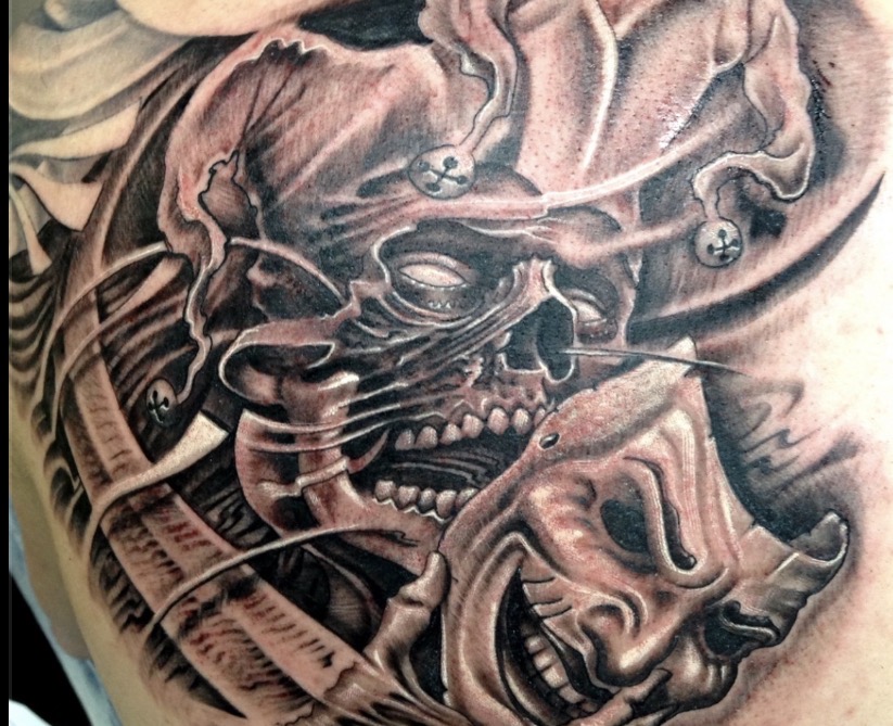 Düsteres Black-and-Grey Tattoo eines Narren (Jester / Joker): Ein Totenkopf mit Hofnarrenkappe und Schellen, der eine lachende Maske zur Seite zieht. Mit surrealen, biomechanischen Strukturen und fließenden Übergängen im Hintergrund.