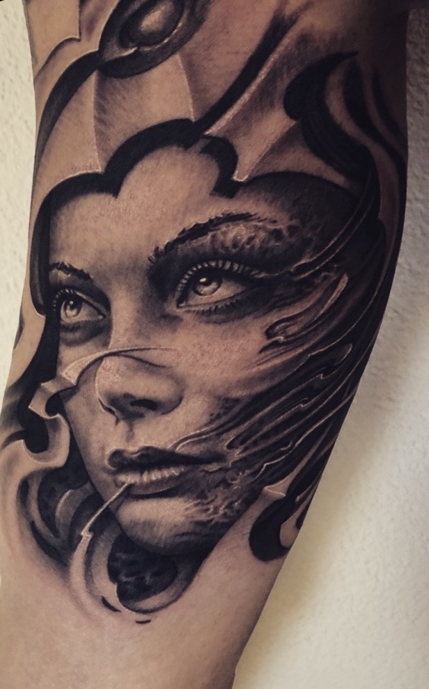 Hochaufgelöstes Black-and-Grey Tattoo auf der Oberarm-Innenseite: Realistic Porträt einer Frau mit intensivem Blick, deren Gesicht auf einer Seite surreal in fließende, schattierte Formen und organisch-biomechanische Elemente auflöst.