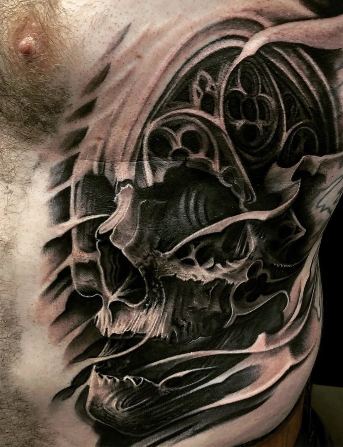 Surreales Black-and-Grey Tattoo im Dark-Art-Stil auf Brust und Rippen: Ein plastischer Totenkopf mit geöffnetem Kiefer, der nahtlos mit gotischen Fensterelementen, filigranem Maßwerk und organisch-fließenden Strukturen verschmilzt.