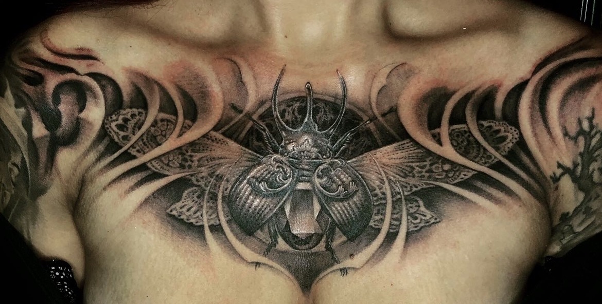 Aufwendiges Black-and-Grey Tattoo auf dem Dekolleté einer Frau: Ein detaillierter Nashornkäfer bzw. Skarabäus mit ausgebreiteten, filigran gemusterten Flügeln, eingebettet in organische, geschwungene Schattierungen und filigrane Spitzen-Ornamentik über der Brust.