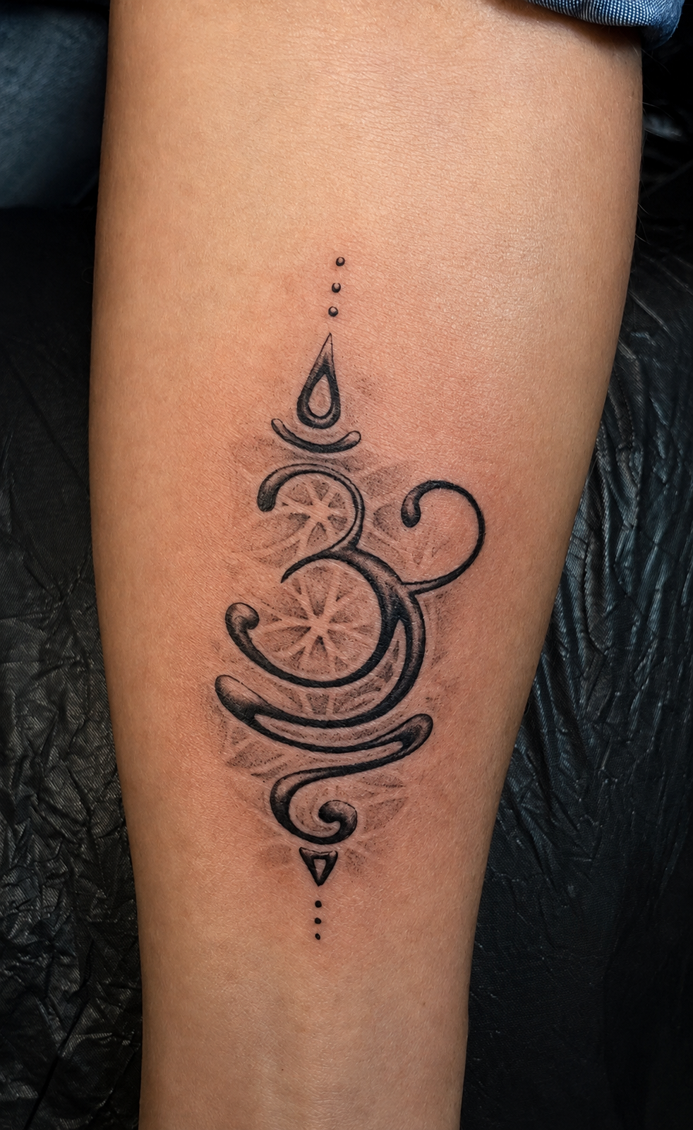 Filigranes Black-and-Grey Tattoo auf dem Arm: Ein elegant geschwungenes Om-Symbol im Unalome-Stil mit feinen Pünktchen-Details, unterlegt mit einer zart schattierten Blume des Lebens (Heilige Geometrie).