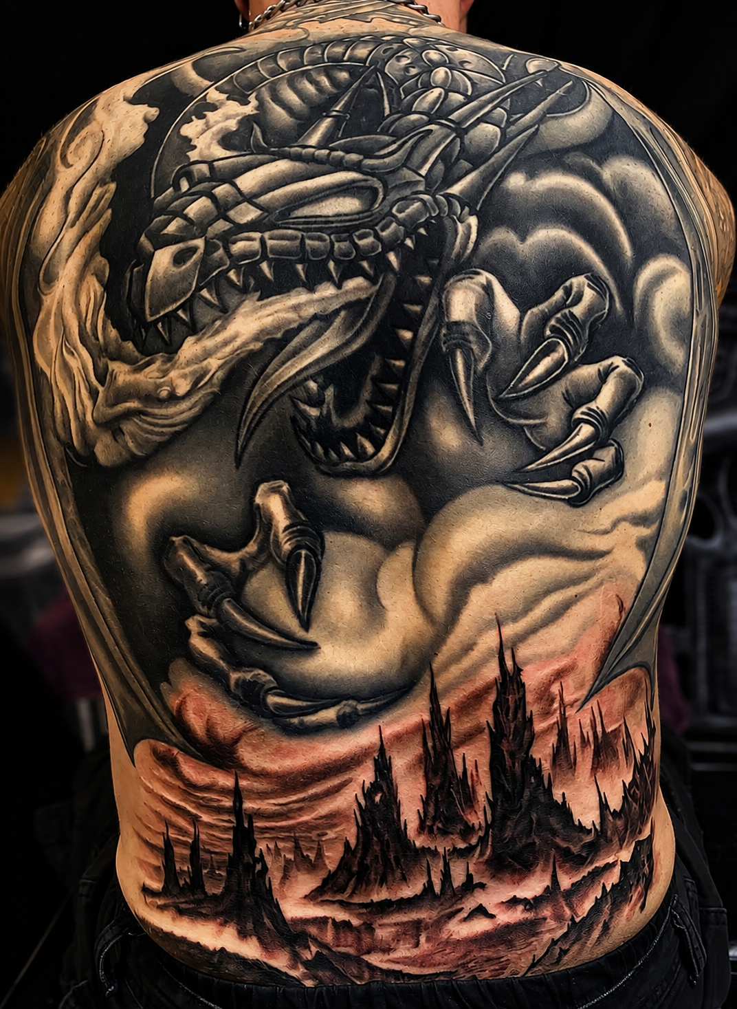 Monumentales Fantasy-Tattoo auf dem gesamten Rücken: Ein mächtiger, feuerspeiender Drache mit spitzen Klauen und gepanzertem Kopf schwebt aus Wolken über einer düsteren, zackigen Berg- und Festungslandschaft in  Grautönen.