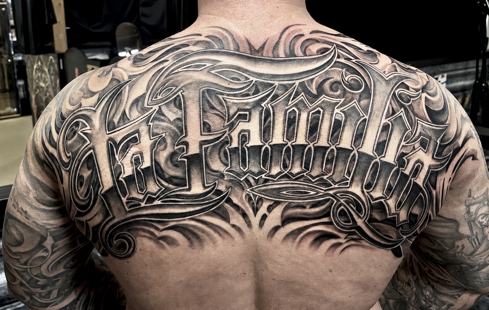 Massives Black-and-Grey Lettering-Tattoo über den gesamten oberen Rücken von Floyd Varesi: Der schwungvolle Schriftzug 'La Familia' in detailliert ausgearbeiteter Custom-Chicano-Schrift, unterlegt mit fließenden, dunklen Filigree-Schattierungen und Schnörkel-Elementen.