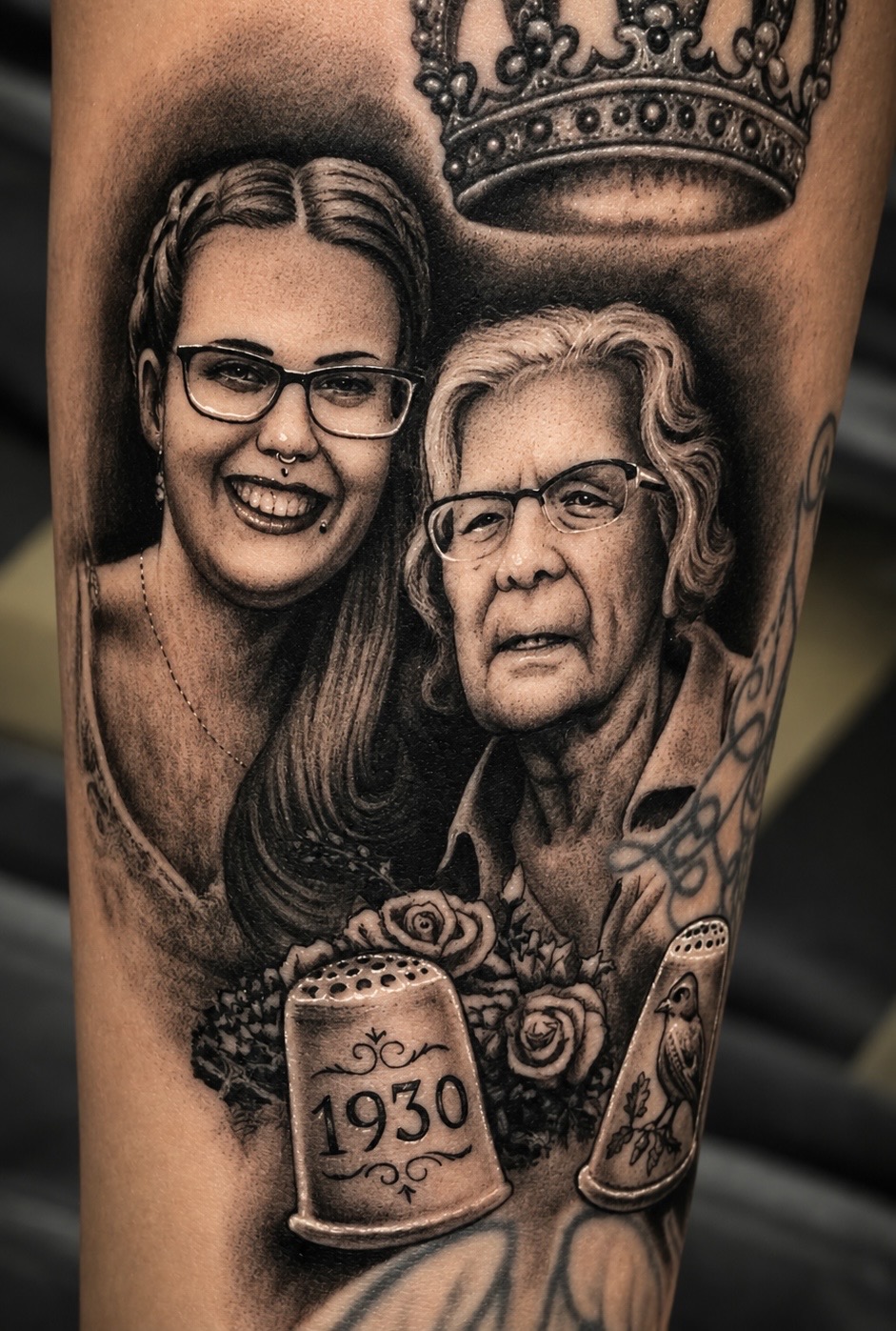 Fein schattiertes Black-and-Grey Porträt-Tattoo auf dem Unterarm: Zwei lächelnde Frauen (eine jüngere und eine ältere Frau mit Brille), darüber eine schwebende Krone. Darunter befinden sich zwei gravierte Fingerhüte mit der Jahreszahl 1930 und einem Vogelmotiv, eingebettet in Rosen.