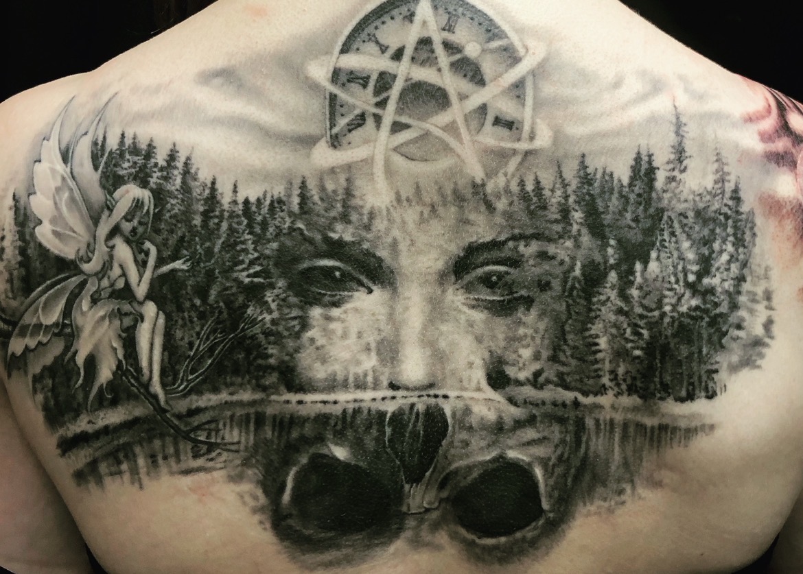 Surreales Black-and-Grey Rücken-Tattoo: Links sitzt eine filigrane Fee auf einem Ast vor einem Nadelwald. In der Mitte verschmilzt ein Frauengesicht mit den Bäumen, dessen Spiegelung im Wasser die optische Täuschung eines Totenkopfs bildet. Darüber schwebt eine Uhr mit Pentagramm- und Atheismus-Symbolik.