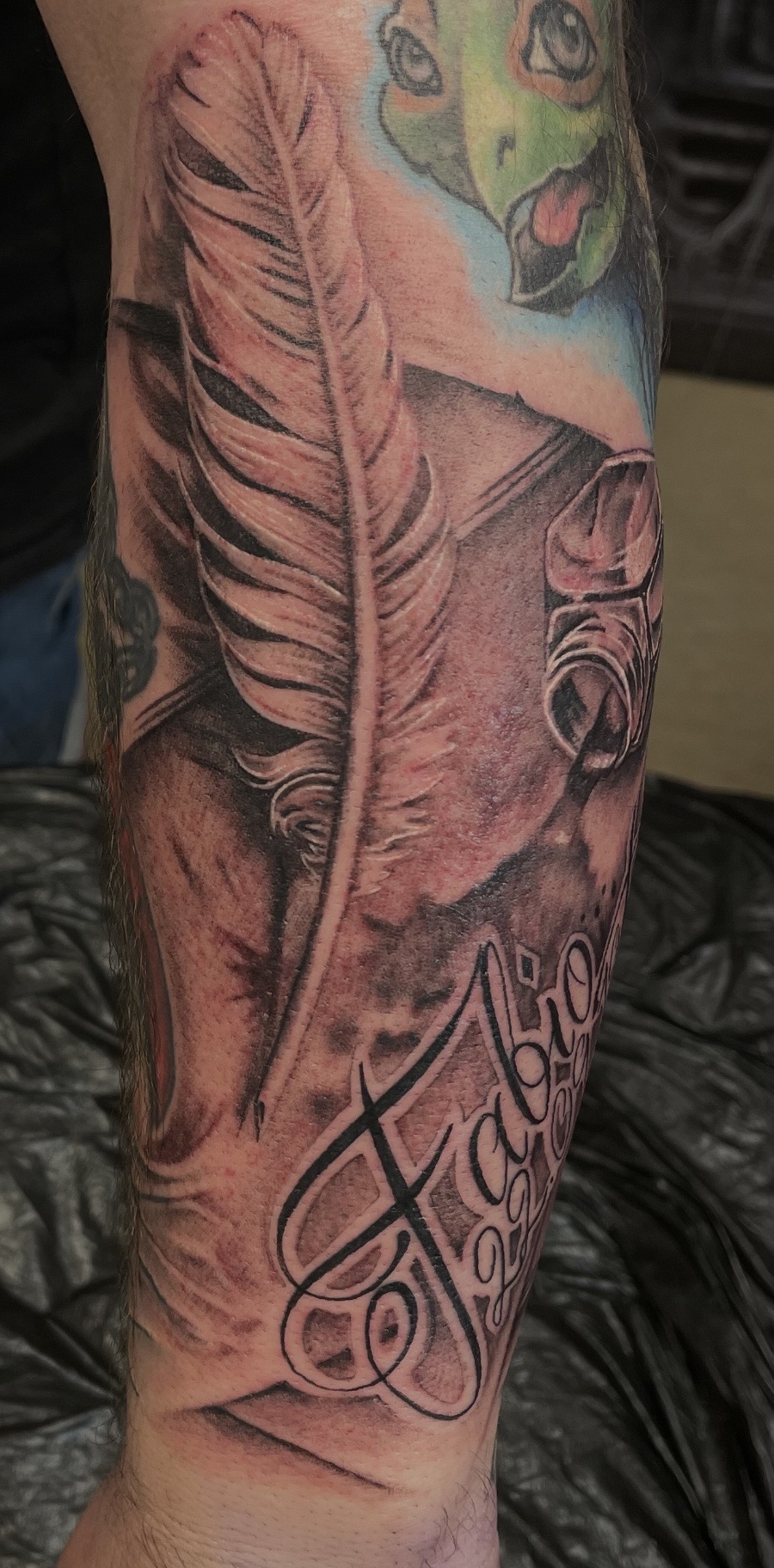 Black-and-Grey Unterarm-Tattoo einer großen, detailreich schattierten Schreibfeder neben einem alten Tintenfass auf geschwungenem Pergamentpapier, verziert mit einem schwungvollen Namenszug ('Fabio').