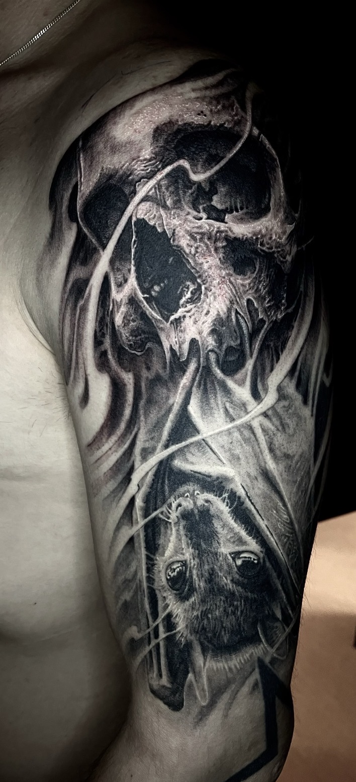 Düsteres Black-and-Grey Dark-Art Tattoo auf dem Oberarm von Tätowierer Floyd Varesi: Oben ein morphinter Totenkopf mit organischen Texturen, darunter eine kopfüber hängende, realistisch ausgearbeitete Fledermaus, eingehüllt in fließende Nebel- und Rauchschleier.