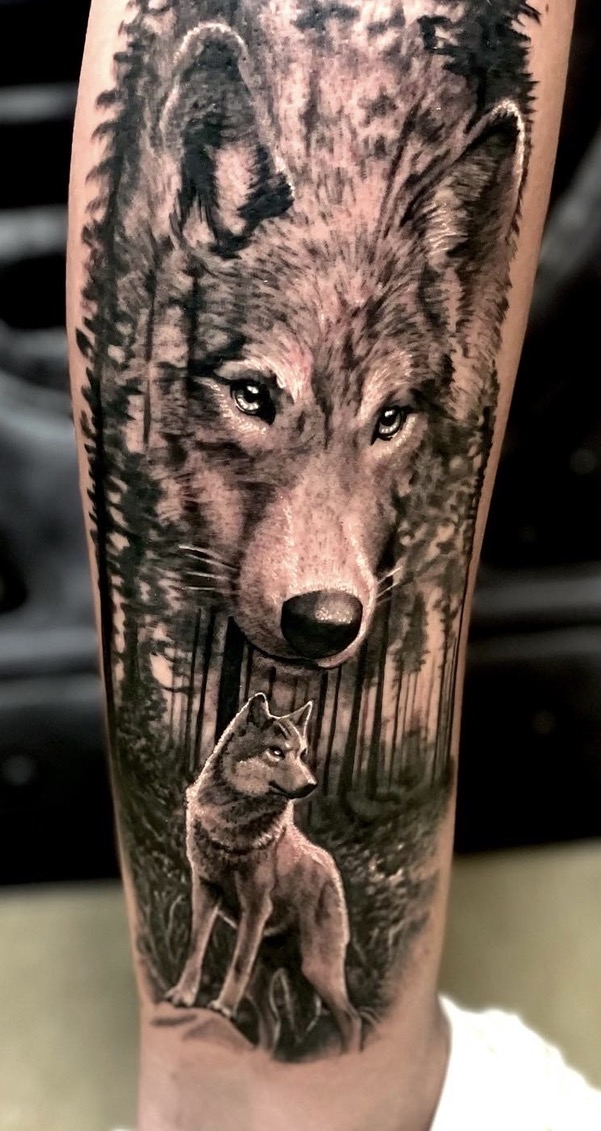 Realistic Black-and-Grey Tattoo auf der Wade/dem Unterschenkel: Ein großer, detailliert ausgearbeiteter Wolfskopf mit klaren Augen, der nahtlos in eine dicht stehende Nadelwald-Landschaft übergeht. Im Vordergrund steht ein kompletter Wolf auf einer Lichtung.