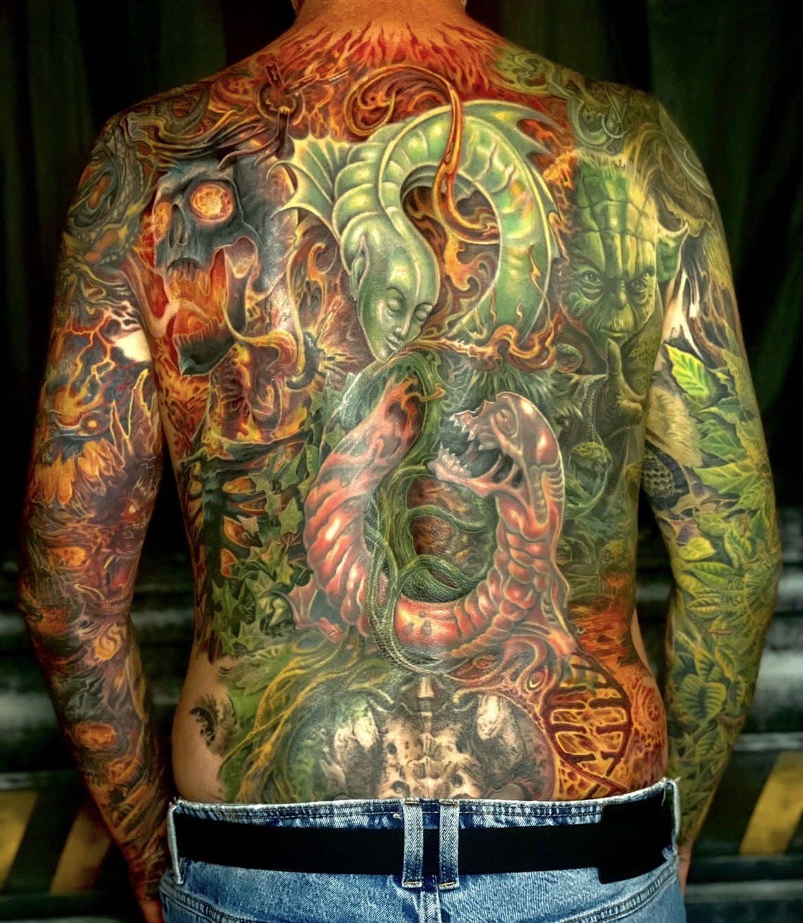Monumentales, farbenprächtiges Rücken-Tattoo zum Thema Gut und Böse, Erschaffen und Vernichten, gemeinsam gestochen von Varry und Floyd Varesi: Sattgrüne Natur- und Myken-Elemente symbolisieren das Leben, während feuriges Rot, brennende Schädel und zerstörerische Kräfte den Gegenpol bilden.